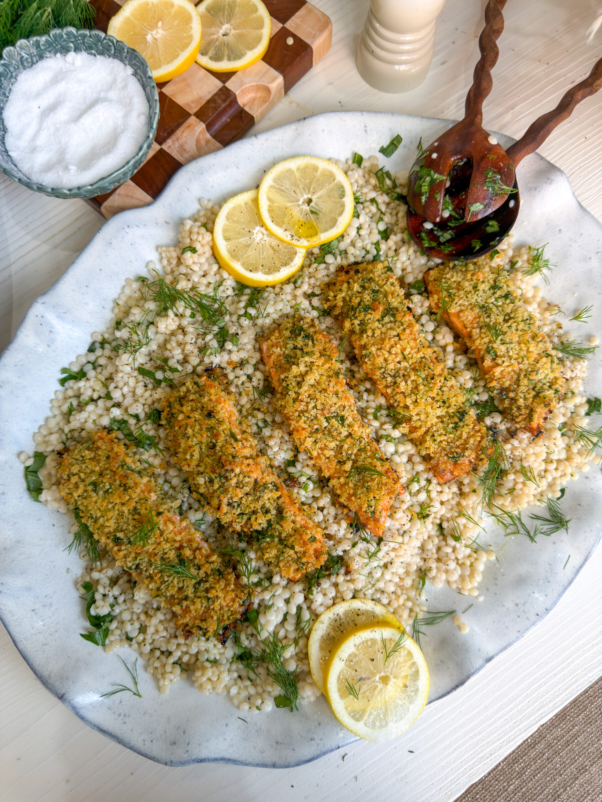 A large platter of lemony pearl couscous topped with five fillets of herb-crusted panko salmon, garnished with fresh dill and lemon slices. Beside it sits a dish of roasted rainbow carrots over a layer of whipped yogurt, sprinkled with pomegranate seeds and herbs. A small bowl of salt, a wooden cutting board with sliced lemons and fresh dill, and other tabletop items surround the dishes on a light wooden surface.