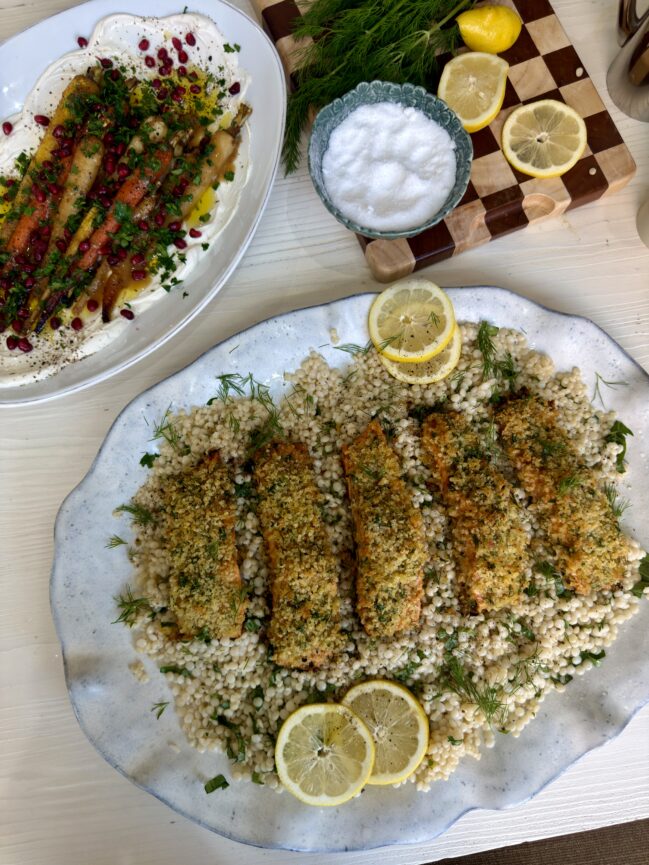 A large platter of lemony pearl couscous topped with five fillets of herb-crusted panko salmon, garnished with fresh dill and lemon slices. Beside it sits a dish of roasted rainbow carrots over a layer of whipped yogurt, sprinkled with pomegranate seeds and herbs. A small bowl of salt, a wooden cutting board with sliced lemons and fresh dill, and other tabletop items surround the dishes on a light wooden surface.