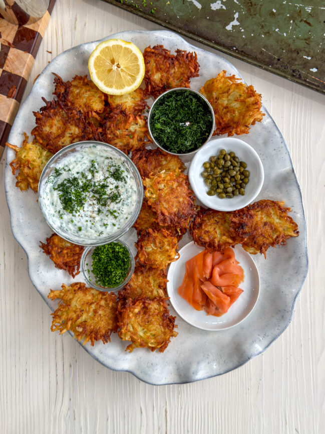 Latke board with crispy potato latkes, dill and chive sour cream dip, smoked salmon, fresh herbs, lemon slices, and capers.”