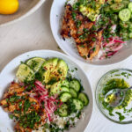 Two white bowls filled with honey-lime cilantro chicken and rice bowls, topped with sliced avocado, cucumbers, pickled red onions, fresh cilantro, lime wedges, and chili flakes. A small bowl of honey-lime dressing sits nearby, and a wooden bowl of lemons is placed beside the plates on a white table.