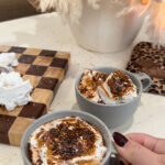 Two gray mugs of rich French hot chocolate topped with toasted homemade marshmallows sit on a light-colored countertop, with a hand holding one mug. A wooden cutting board with marshmallow pieces and a neutral dried floral arrangement are in the background, creating a cozy, café-style setting.