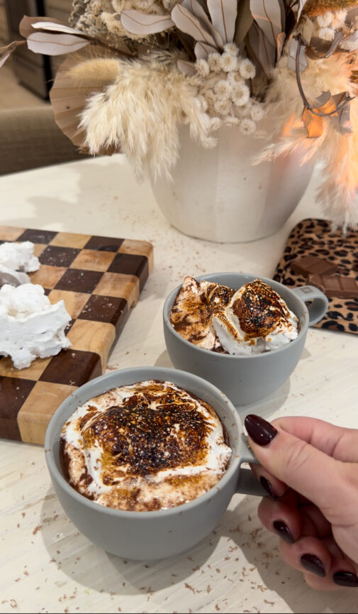 Two gray mugs of rich French hot chocolate topped with toasted homemade marshmallows sit on a light-colored countertop, with a hand holding one mug. A wooden cutting board with marshmallow pieces and a neutral dried floral arrangement are in the background, creating a cozy, cafรฉ-style setting.