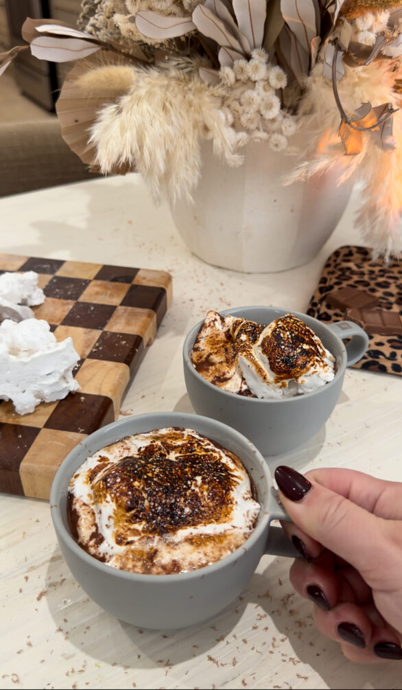 Two gray mugs of rich French hot chocolate topped with toasted homemade marshmallows sit on a light-colored countertop, with a hand holding one mug. A wooden cutting board with marshmallow pieces and a neutral dried floral arrangement are in the background, creating a cozy, café-style setting.